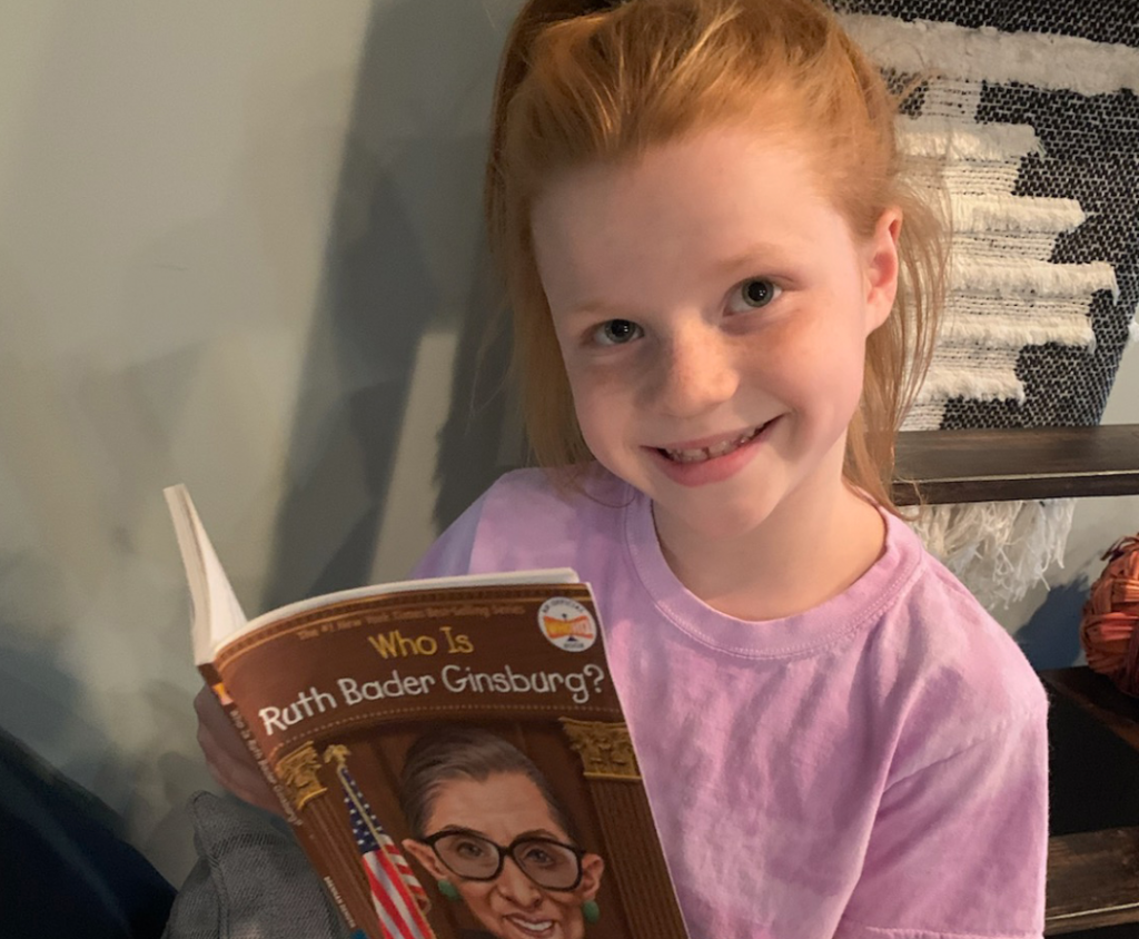 Smiling young girl with red hair and freckles, holding a book titled, "Who is Ruth Bader Ginsburg?"