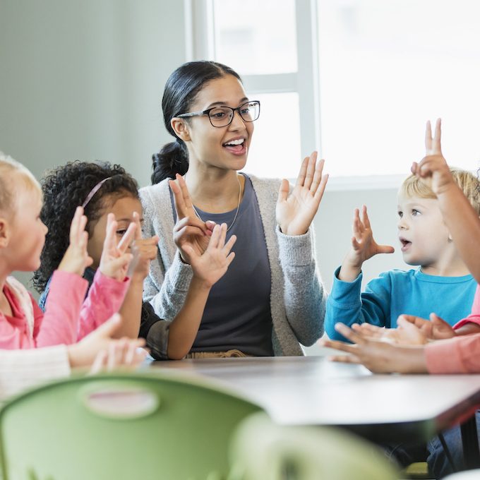 A multi-ethnic group of six preschool children with a mixed race African-American and Caucasian teacher, sitting around a table in a classroom. The teacher and some of her students have their hands raised, holding up fingers, learning how to count. The children are 4 years old.